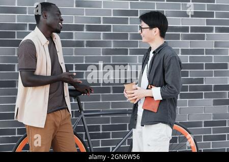 Horizontal side view shot of handsome African American guy and stylish Asian woman standing together outdoors enjoying conversation Stock Photo