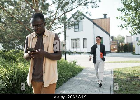 Horizontal shot of young African American man using his smartphone while walking out on summer day, confident Asian woman going to somewhere Stock Photo