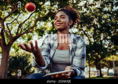 Mixed race female holding digital tablet researching project on healthy foods while playing with fresh red tomato Stock Photo