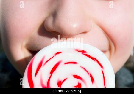 A large round lollipop held close to a child's mouth. Clearly the child is happy even if the face is not entirely visible. Sunny natural day light. Stock Photo
