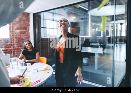 Two creative businesswomen sticking adhesive notes to a glass wall with their colleagues in the background. Two focused young businesswomen sharing th Stock Photo