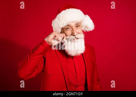 Photo of handsome imposing santa claus cheerful beaming smile play mustache wear x-mas hat suit on red color background Stock Photo