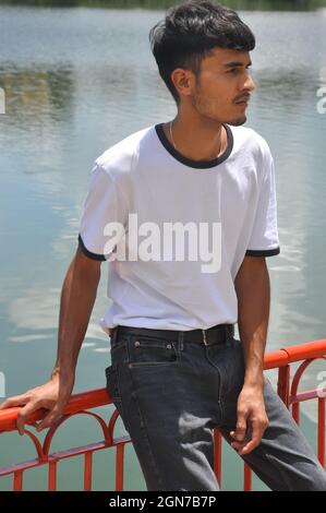 A good looking Indian young guy wearing white t-shirt and black jeans with sitting on safety barrier by lake and looking sideways Stock Photo