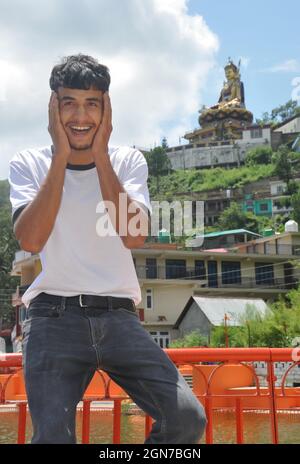 A young guy posing funny sitting on safety barrier beside of lake with beautiful hilly area of Tso-Pema and statue of Padmasambhava (Guru Rinpoche) in background Stock Photo