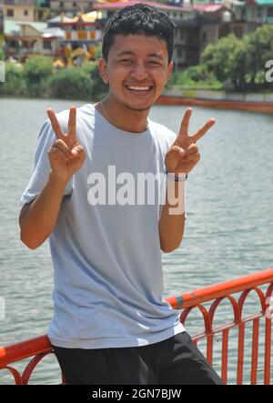 A Ladakhi young guy smiling sitting on safety barrier by lake with making victory hand sign and looking at camera Stock Photo