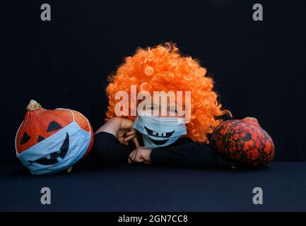 boy 4 years old in orange wig and pumpkin in blue medical mask with a painted scary smile. On black background. Halloween decorations and celebrations Stock Photo