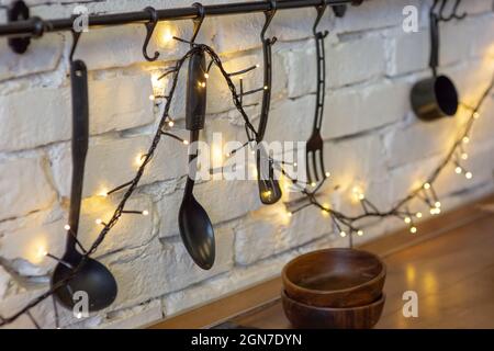 Black kitchen utensils hang on a white brick wall, decorated with glowing garlands Stock Photo