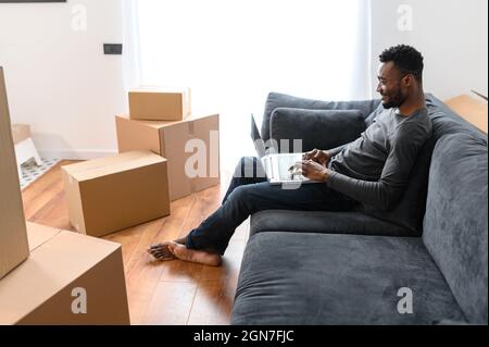 Side view at satisfied African-American guy sitting on the comfortable couch and using laptop computer among cardboard boxes, smiling black man moving into new apartment, shopping online, web surfing Stock Photo
