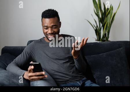 Surprised African-American guy sitting on the couch and holding smartphone, staring at the screen amazed Stock Photo