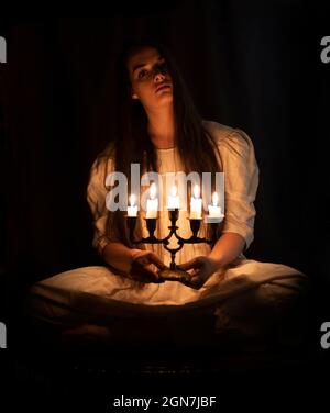 A young girl in an old white dress sitting and holding a candleabra in her hands. Dark background. Scary horror concept. Stock Photo