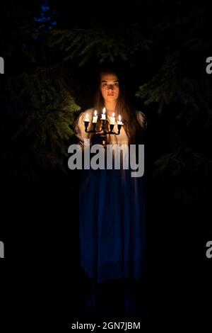 A young woman in the dark woods holding a candelabrum. Wearing an old long white dress. Stock Photo
