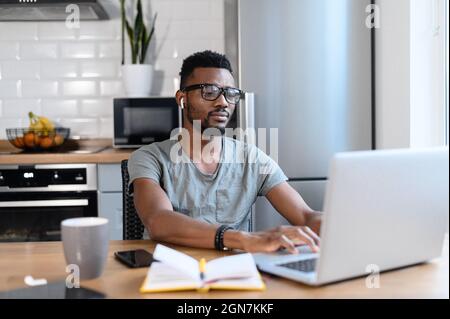 Concentrated African-American guy is working with a laptop sitting in the kitchen. Black man in casual wear and eyeglasses is typing on the keyboard, answering email, communicates with the customer Stock Photo