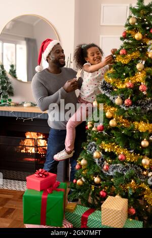 Happy african american father wearing santa hat and daughter decorating christmas tree. christmas, festivity and tradition at home. Stock Photo