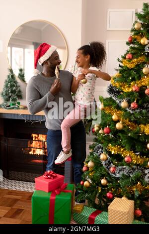 Happy african american father wearing santa hat and daughter decorating christmas tree. christmas, festivity and tradition at home. Stock Photo