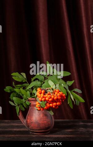 Rowan branch with berries in a clay jug on dark wooden background and textile with copy space Stock Photo