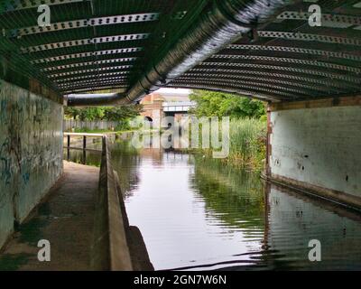 The darkened towpath of the Leeds Liverpool Canal as it passes under a rivetted metal bridge and pipework at the A5057. Stock Photo