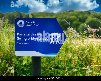 A blue Canal and River Trust sign on a canal towpath with white lettering advises those in rowing boats and canoes to carry their boat around a bridge Stock Photo