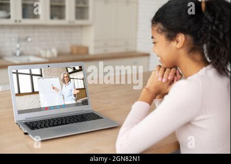 E-studying concept. An African-American female student watching online educational webinar, listening a senior teacher with whiteboard on the screen, preparing to exams online Stock Photo