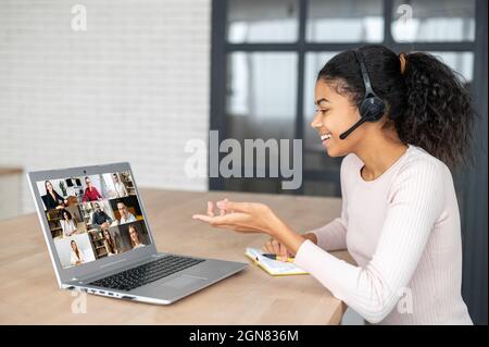 Virtual video conference, online meeting with a many employees together. A young African woman wearing headset is communicating via video call with coworkers, many people on the laptop screen Stock Photo