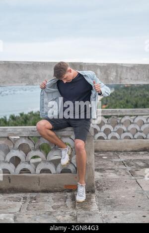 Young handsome Caucasian man in casual clothes and denim jacket, sitting and looking down. Sitting on the roof and holding his jacket Stock Photo
