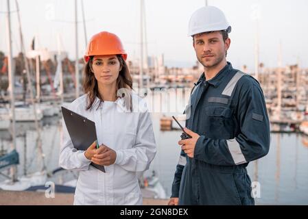 Serious young male and female colleagues in hardhats and protective uniform with documents and walkie talkie looking at camera while inspecting sea port together Stock Photo