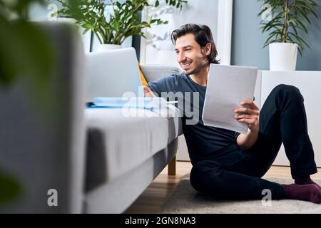 Male freelance worker holding documents while working on laptop at home Stock Photo