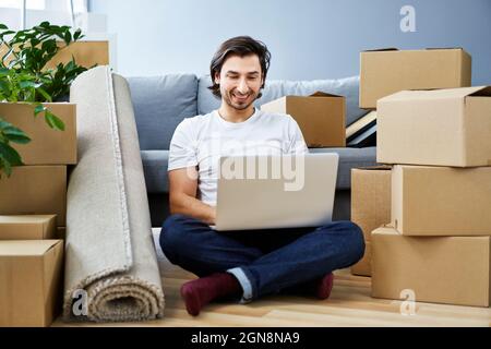 Smiling man using laptop while sitting amidst cardboard boxes at home Stock Photo