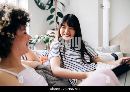 Happy woman with digital tablet looking at friend while sitting on sofa Stock Photo
