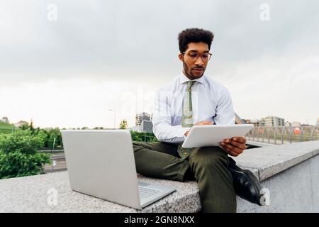 Male business professional using digital tablet while sitting with laptop on retaining wall Stock Photo