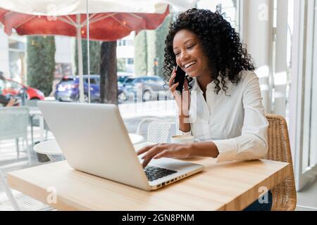 Businesswoman talking on mobile phone while using laptop at coffee shop Stock Photo
