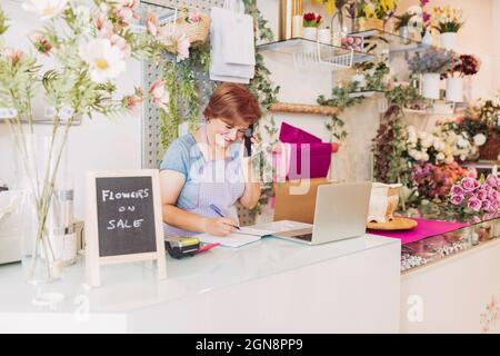 Smiling female flower shop owner talking on smart phone while writing in diary at checkout Stock Photo