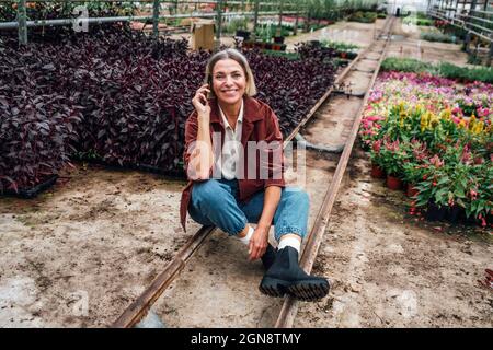 Smiling female farmer talking on mobile phone while sitting in plant nursery Stock Photo