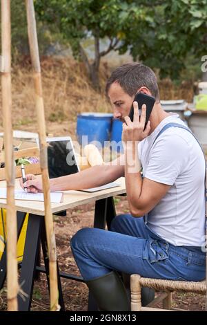Farm worker talking on mobile phone while writing on clipboard sitting at table Stock Photo