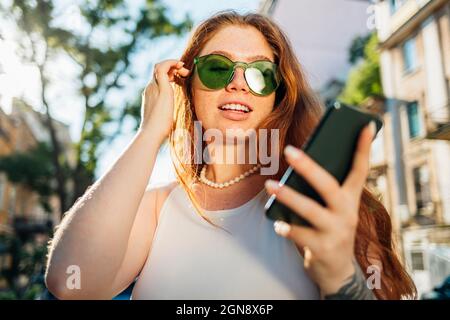 Young woman with hand in hair wearing green sunglasses using smart phone Stock Photo