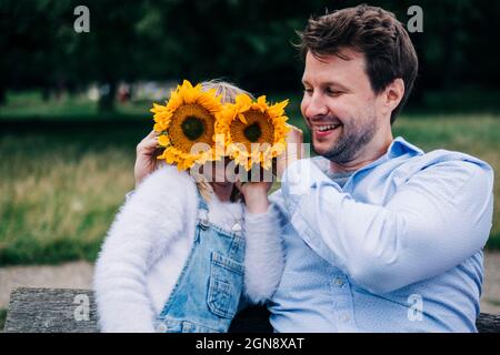 Smiling man covering girl's face with sunflowers while sitting on bench Stock Photo