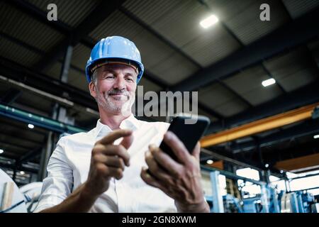 Smiling male engineer with hardhat using mobile phone at metal industry Stock Photo