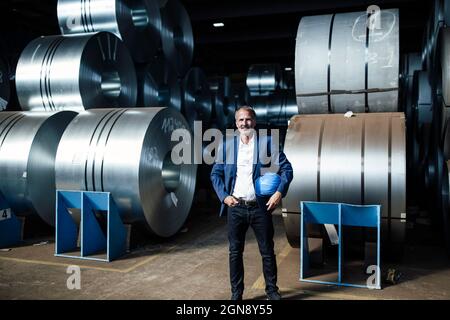 Male professional holding hardhat while standing with hand in pocket at warehouse Stock Photo