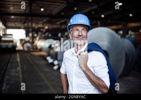 Cheerful male engineer with hardhat holding blazer at warehouse Stock Photo