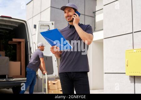 Smiling delivery man holding clipboard while talking on smart phone Stock Photo