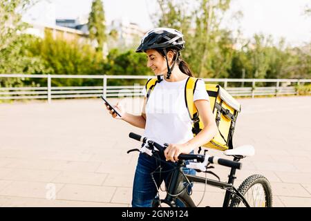Smiling delivery woman using mobile phone while wheeling bicycle Stock Photo