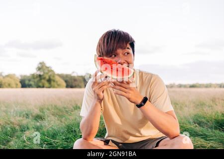 Young man holding watermelon slice while sitting on meadow Stock Photo