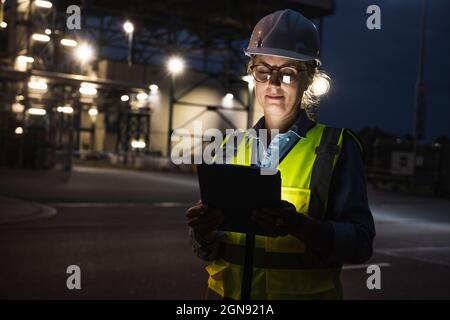 Female engineer with hardhat using digital tablet outside factory at night Stock Photo