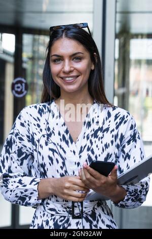Smiling young businesswoman holding mobile phone and laptop Stock Photo