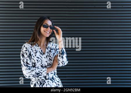 Young woman smiling while wearing sunglasses in front of black wall Stock Photo