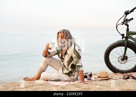 Man with long gray hair holding mug while sitting by bicycle at beach Stock Photo