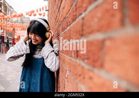 Cheerful young woman wearing headphones by brick wall Stock Photo