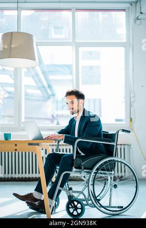 Young disabled male professional using laptop while sitting in wheelchair in office Stock Photo