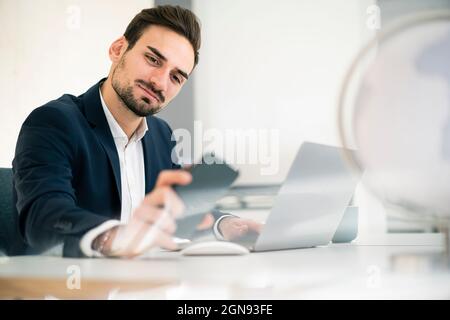 Handsome male professional checking smart phone while sitting with laptop at desk in office Stock Photo