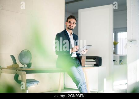 Handsome young businessman holding digital tablet while sitting on desk in office Stock Photo