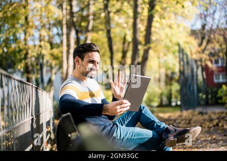 Young man waving hand while doing video call through digital tablet at park during sunny day Stock Photo